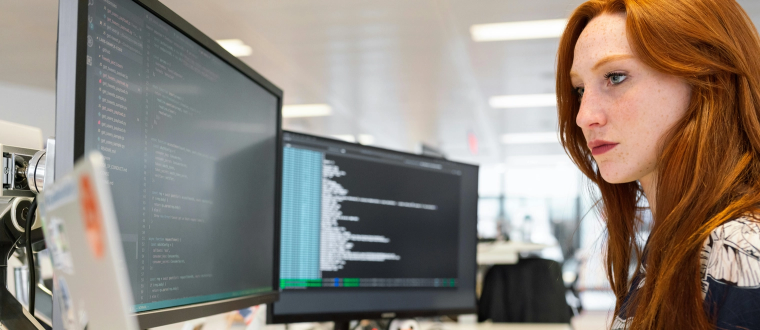 Woman coding on dual computer monitors in office.